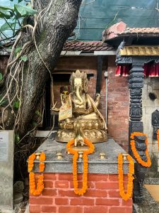 A golden statue of the Hindu god Ganesha sits on a brick platform draped with bright orange marigold garlands. A large tree trunk is on the left.