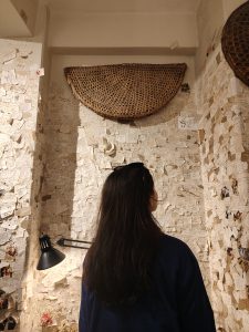 

A person with long dark hair stands in a room where walls are covered with notes and clippings, lit by a black lamp beneath a woven wall basket.