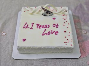A white square cake with the words "41 Years of Love" written in pink icing. Decorated with red candy pearls and ribbon-shaped white chocolate, placed on a light tablecloth, celebrating a long-lasting love story. 