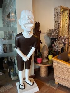 A large, white statue of a laughing man with his hands clasped is wearing a brown shirt and shorts, standing in a decorated indoor area next to a wicker chair and a small table with potted plants.