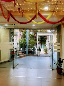 The entrance to a building with open glass doors and a light gray tile floor. The ceiling is decorated with red and yellow fabrics and string lights, leading out to an exterior staircase and courtyard.