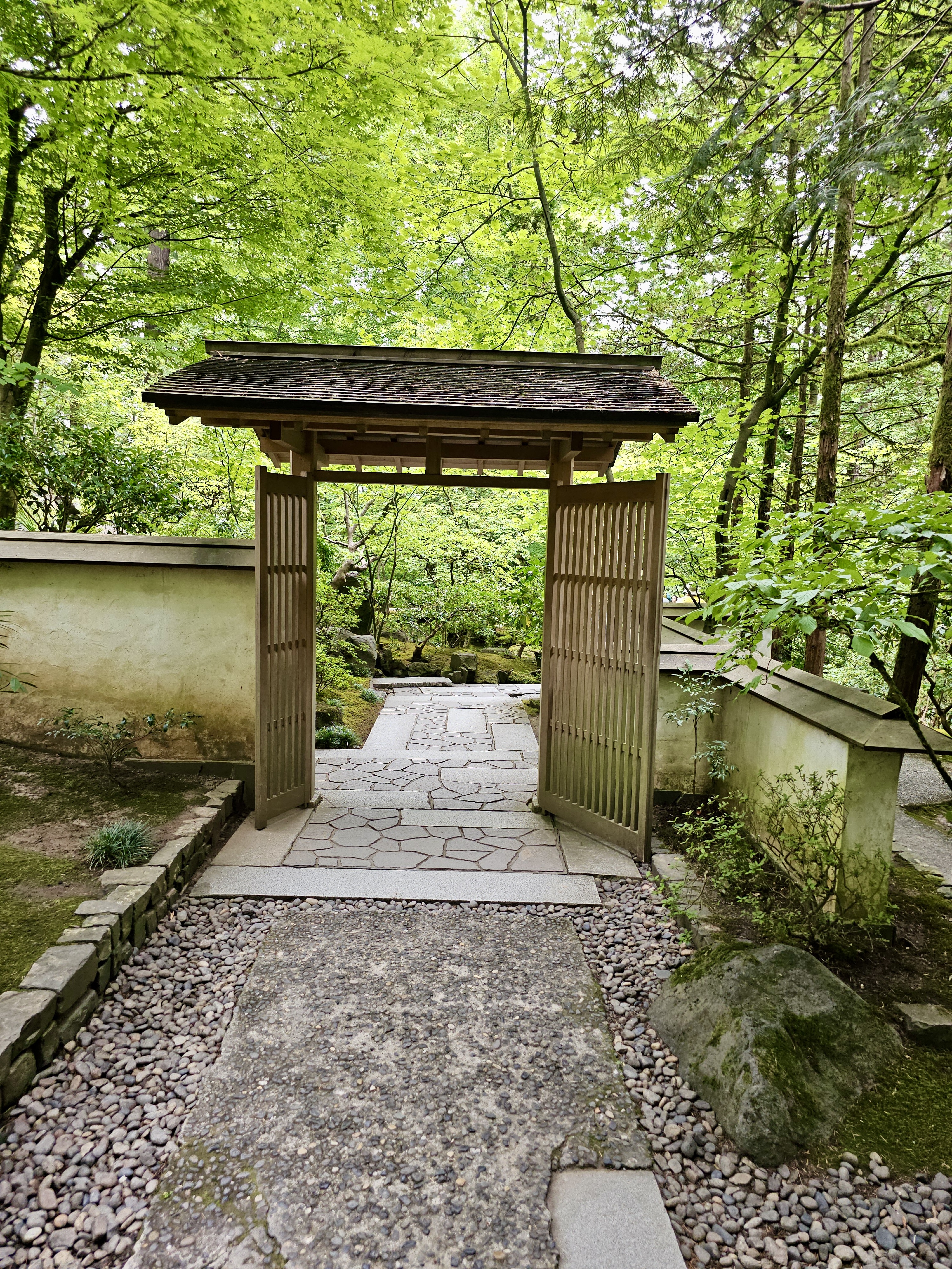 An open wooden gate leads into a peaceful garden path surrounded by tall trees and stone walls at the Portland Japanese Garden. 