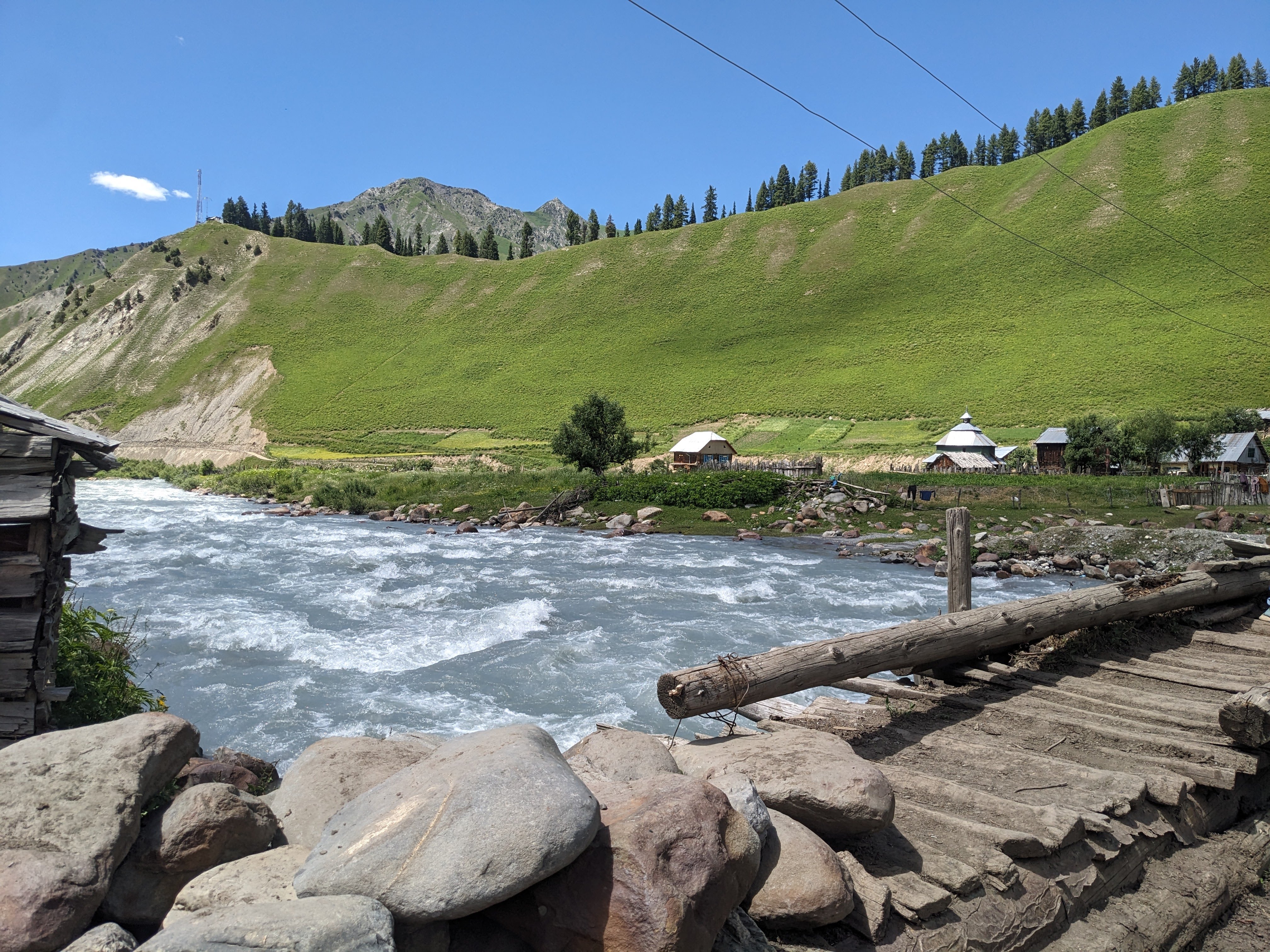 A scenic view of a river flowing swiftly past a rocky shoreline, with green hills in the background dotted with trees. 