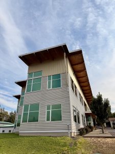 Modern three-story building with green-tinted windows and a sloped roof, surrounded by grass and sidewalk, captured from the streets of Portland. 