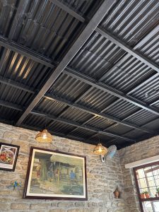 Corrugated brown metal ceiling with exposed frame, tops a stone interior walled room, two wicker basket shaded lit lights hang from the ceiling. A retro wall mounted electric fan sits below them. Window to the right, with small pot plants on the centre frame, two pictures on the stone wall and along with pottery bird ornament.