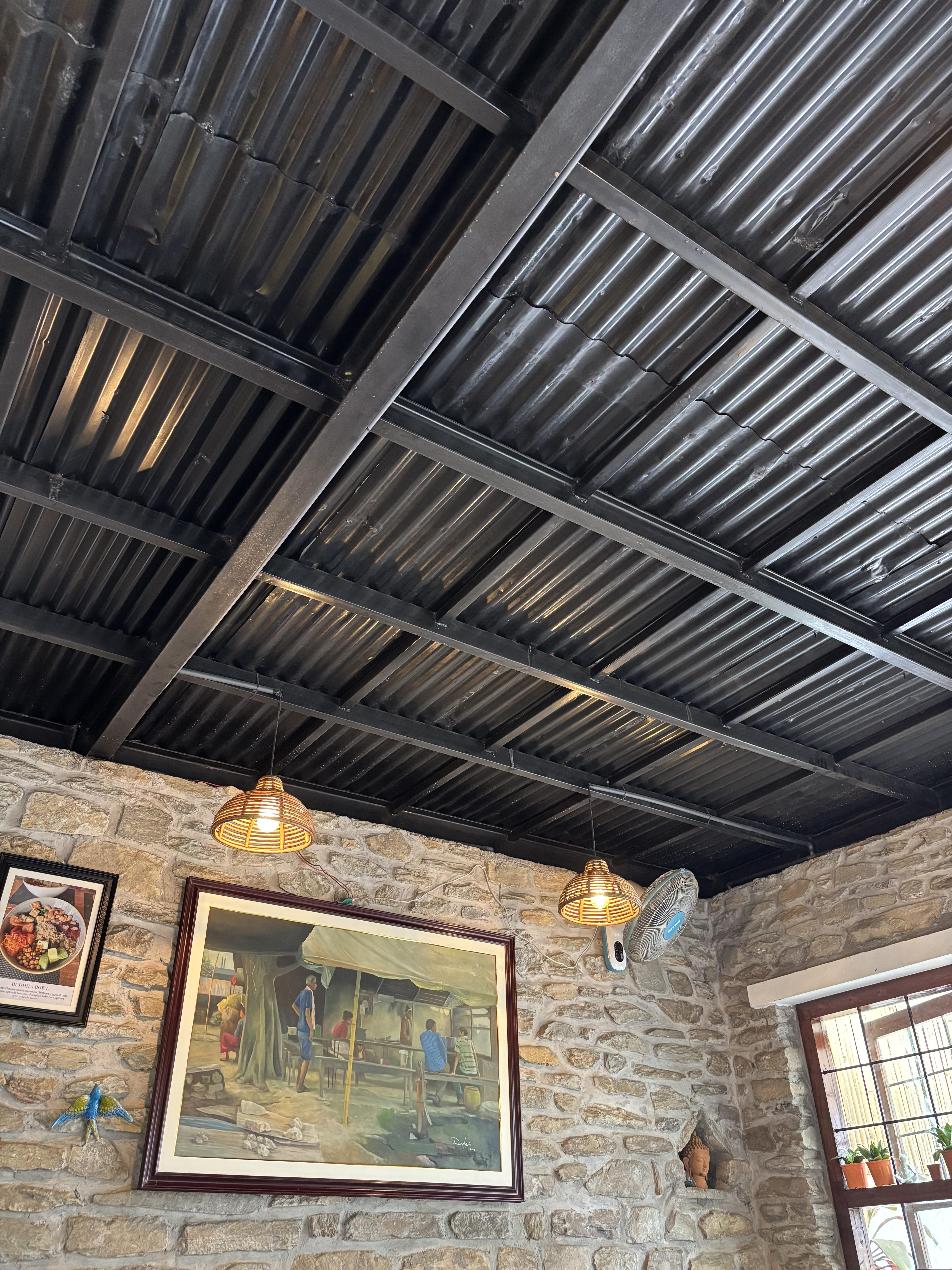 Corrugated brown metal ceiling with exposed frame, tops a stone interior walled room, two wicker basket shaded lit lights hang from the ceiling. A retro wall mounted electric fan sits below them. Window to the right, with small pot plants on the centre frame, two pictures on the stone wall and along with pottery bird ornament.