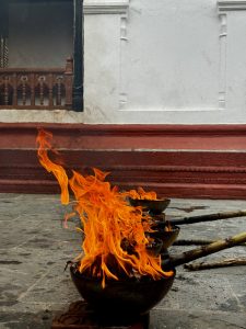 Bowls filled with vibrant flames rest on stone pavement, contrasted against a red and white temple wall, evoking a serene and spiritual ambiance. Also, Lakh Batti is a popular Hindu ritual in Nepal and India where people light 100,000 small wick lamps. It’s usually done as a prayer or promise to a deity, often asking for a wish to come true.