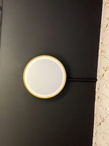 A close-up shot of a small, modern-looking, circular light fixture with a white face and a yellow rim, mounted on a dark surface.