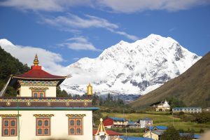 A scenic view featuring a colorful, ornate building with a red and gold roof in the foreground, against a backdrop of snow-capped mountains and a clear blue sky. 