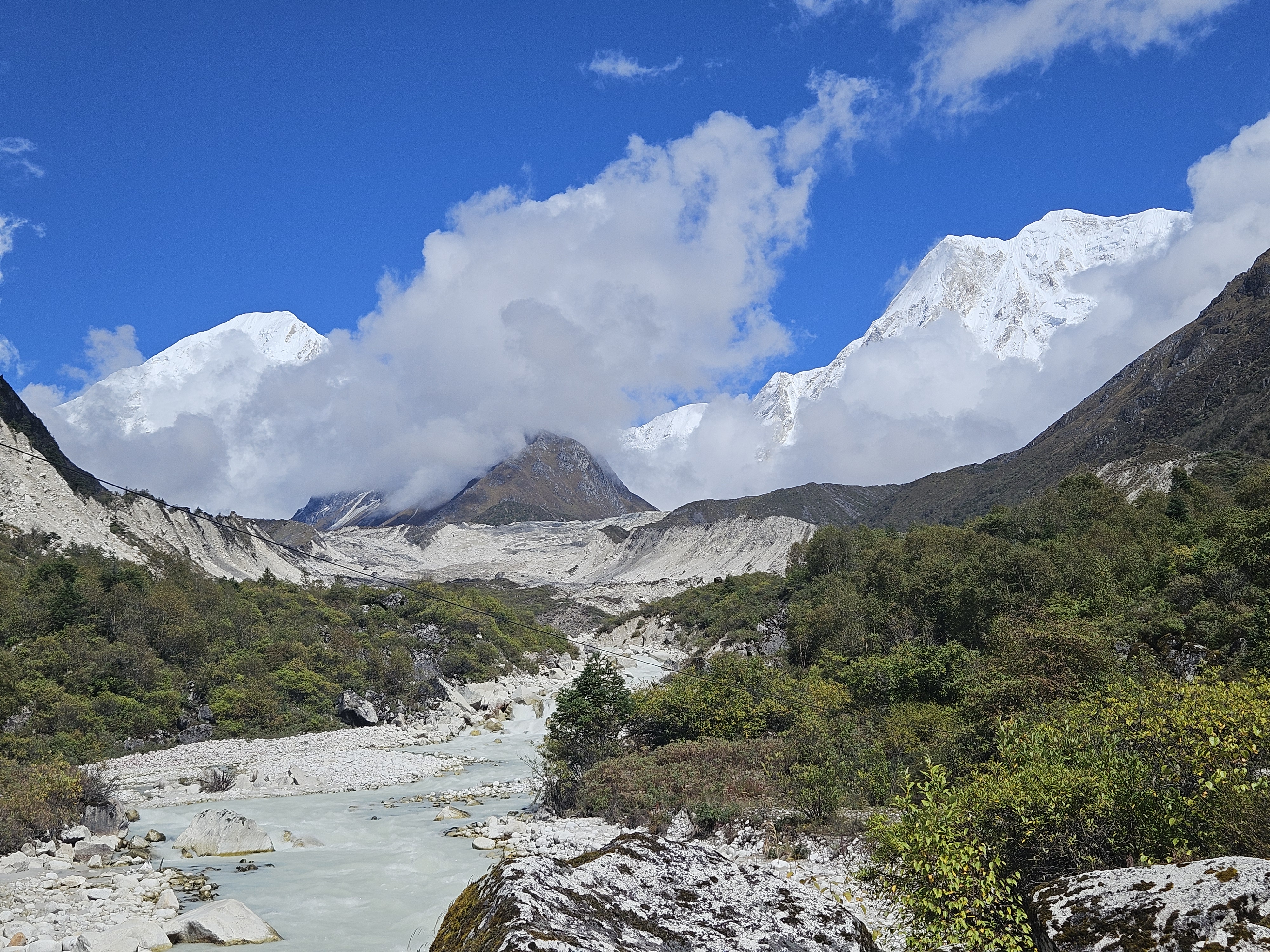 A stunning mountainous landscape featuring towering snow-capped peaks under a bright blue sky, partially obscured by clouds.