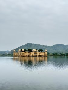 A serene view of Jal Mahal, a historical palace in Jaipur, Rajasthan, surrounded by calm lake waters with lush Aravalli hills in the background under a cloudy sky.