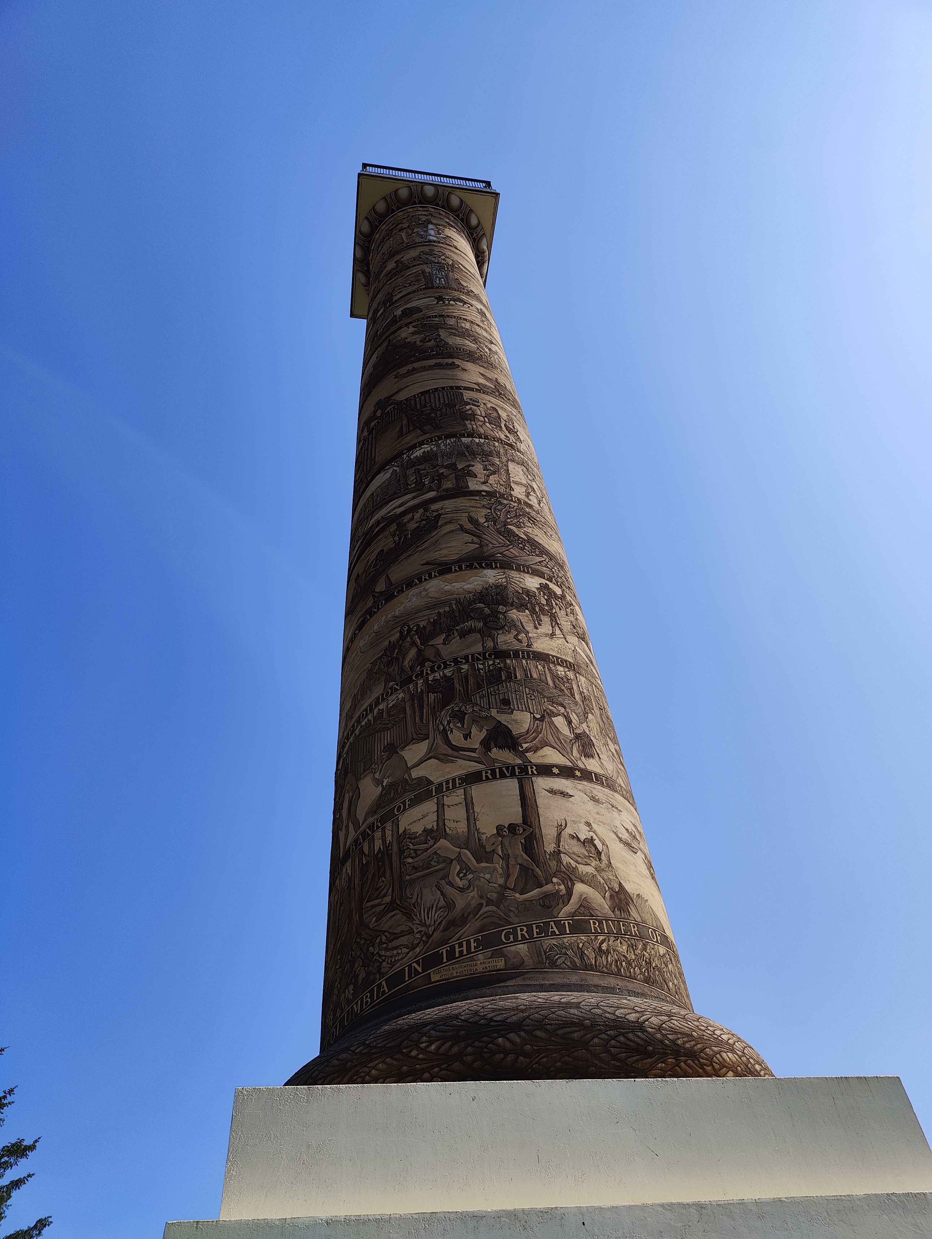 An image of the Astoria column, a historic monument. A tall cylindrical structure with a spiral frieze wrapping around it, behind is the clear blue sky.