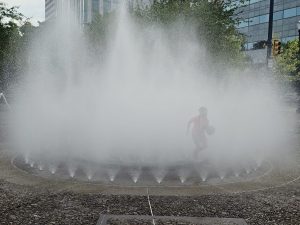 A child runs with a ball through the water jets of a circular fountain at Salmon Street Springs in Riverfront Portland. The mist creates a playful and magical scene. 