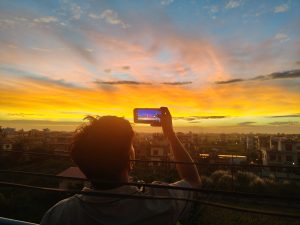 A person stands on a balcony, holding a smartphone up to capture a breathtaking sunset.
