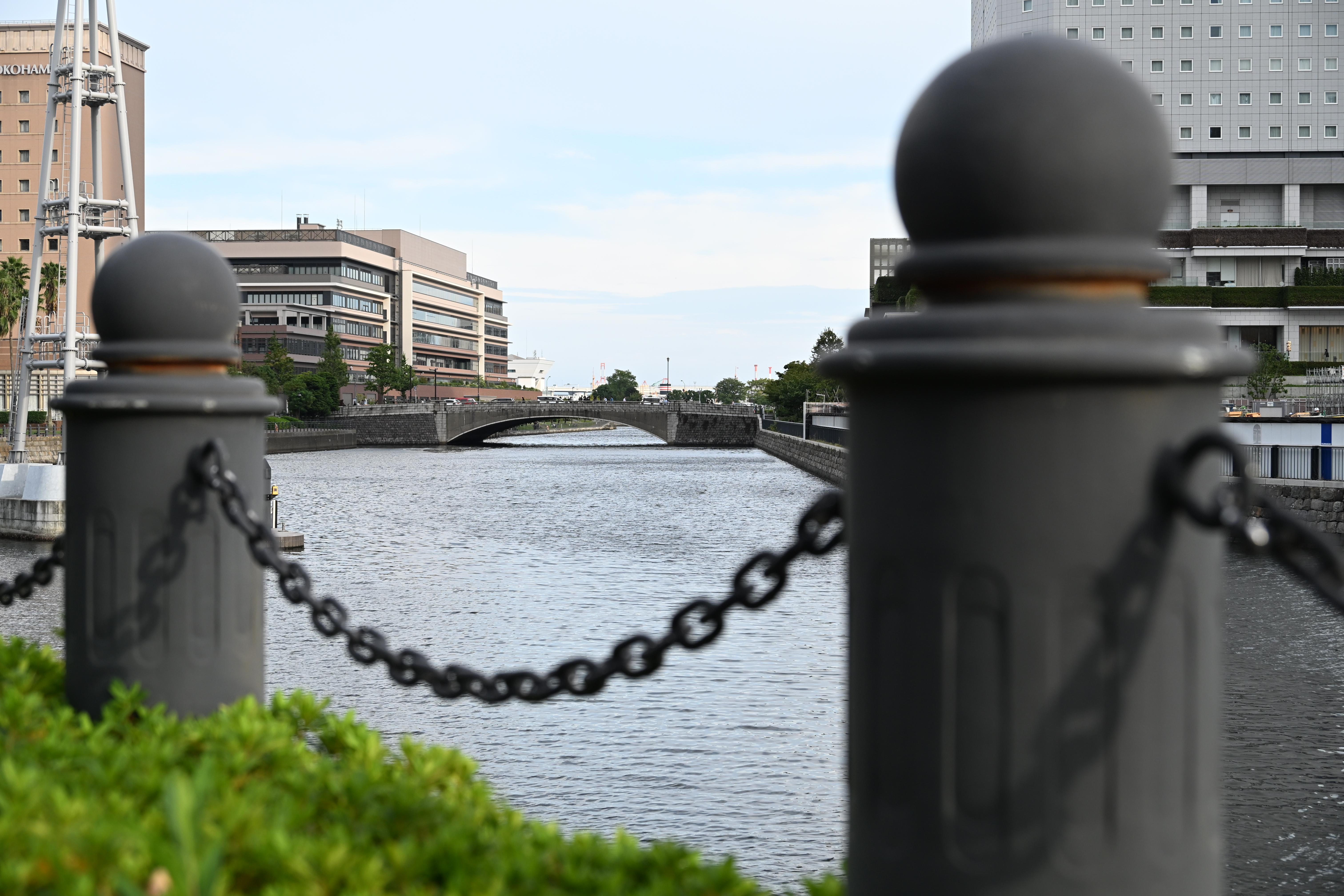 A scenic view of a waterway bordered by a chain-link fence with dark, round posts in the foreground, with modern buildings lining the bank and a bridge spanning the water in the background.