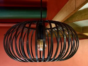 A black metal pendant light fixture with a round cage design hangs from the ceiling of a cafe in Palazhi, Kozhikode. The red wall and wooden beam add a warm, rustic background. 