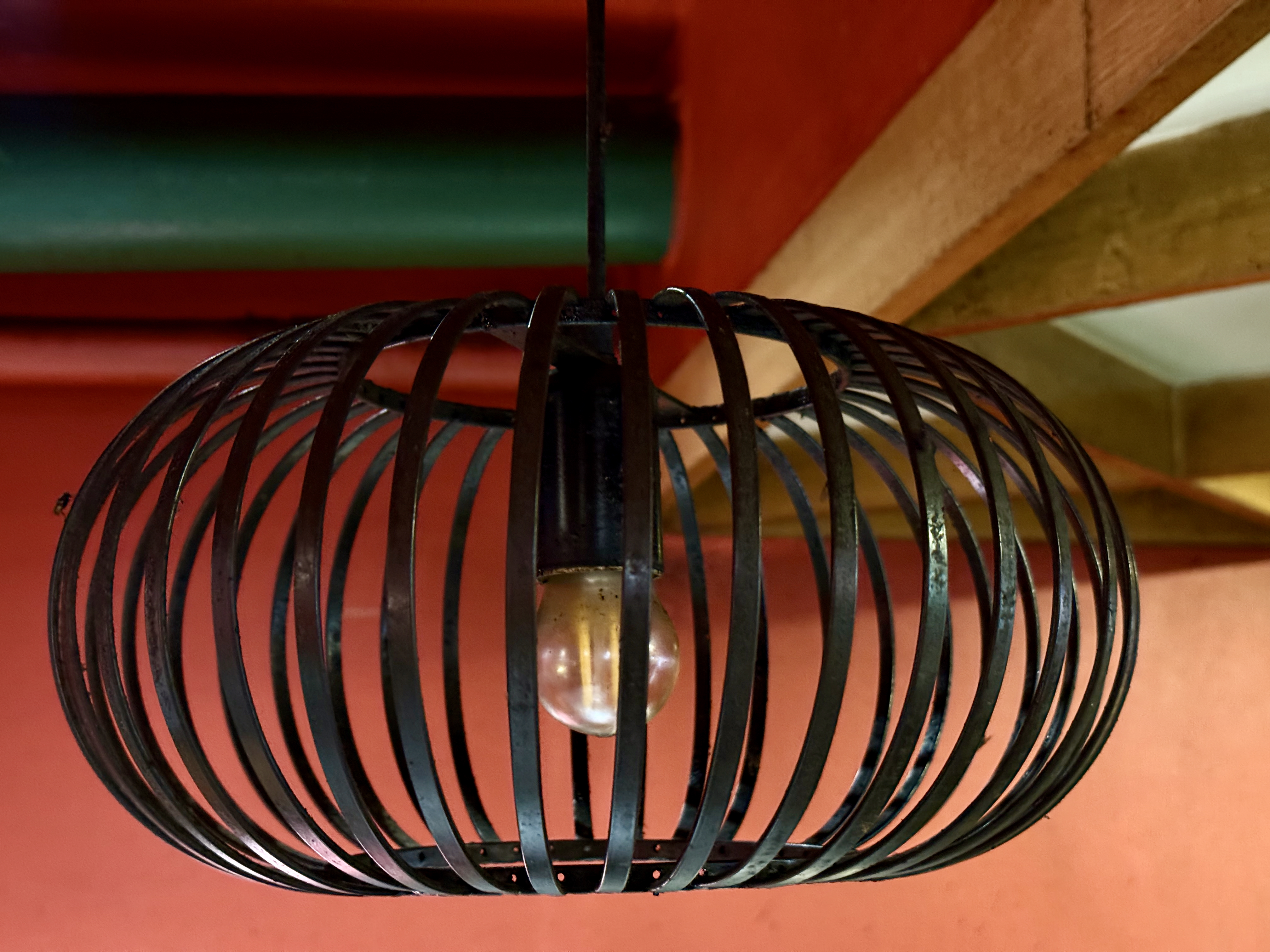A black metal pendant light fixture with a round cage design hangs from the ceiling of a cafe in Palazhi, Kozhikode. The red wall and wooden beam add a warm, rustic background.