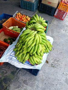 A pile of green bananas rests on a blue crate covered with a white sheet, surrounded by colorful crates of tomatoes, cucumbers, and bell peppers in an open-air market.