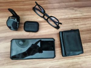 A flat lay of everyday items on a wooden surface, including a smartwatch, a pair of black eyeglasses, a black wireless earbud case, a smartphone displaying a hand reflection on its screen, and a black wallet.
