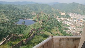 Aerial view of the historic Amer Fort, showing ancient fortification walls winding through green hills, with a water reservoir and the town of Jaipur, Rajasthan.
