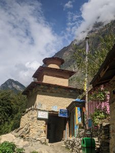 A small, traditional building made of stone and mud sits at the foot of a steep, mountainous terrain under a partly cloudy sky. 