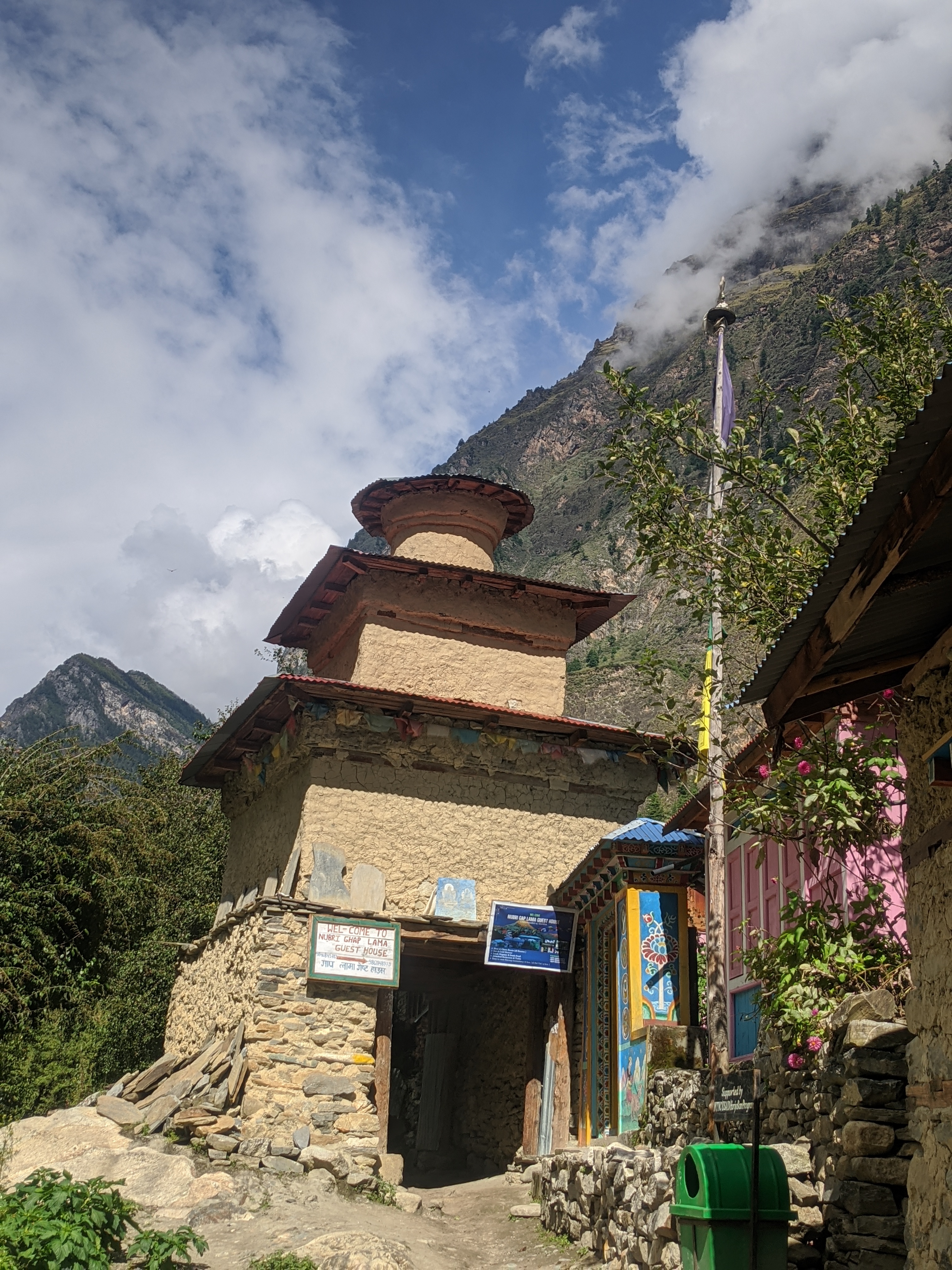 A small, traditional building made of stone and mud sits at the foot of a steep, mountainous terrain under a partly cloudy sky. 