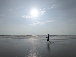 A bright sunny moment at Lal Kakra Beach, Cox’s Bazar. The calm sea glistens under the golden sunlight as a boy walks along the shore with a ball, capturing the simple joy and peaceful rhythm of coastal life.