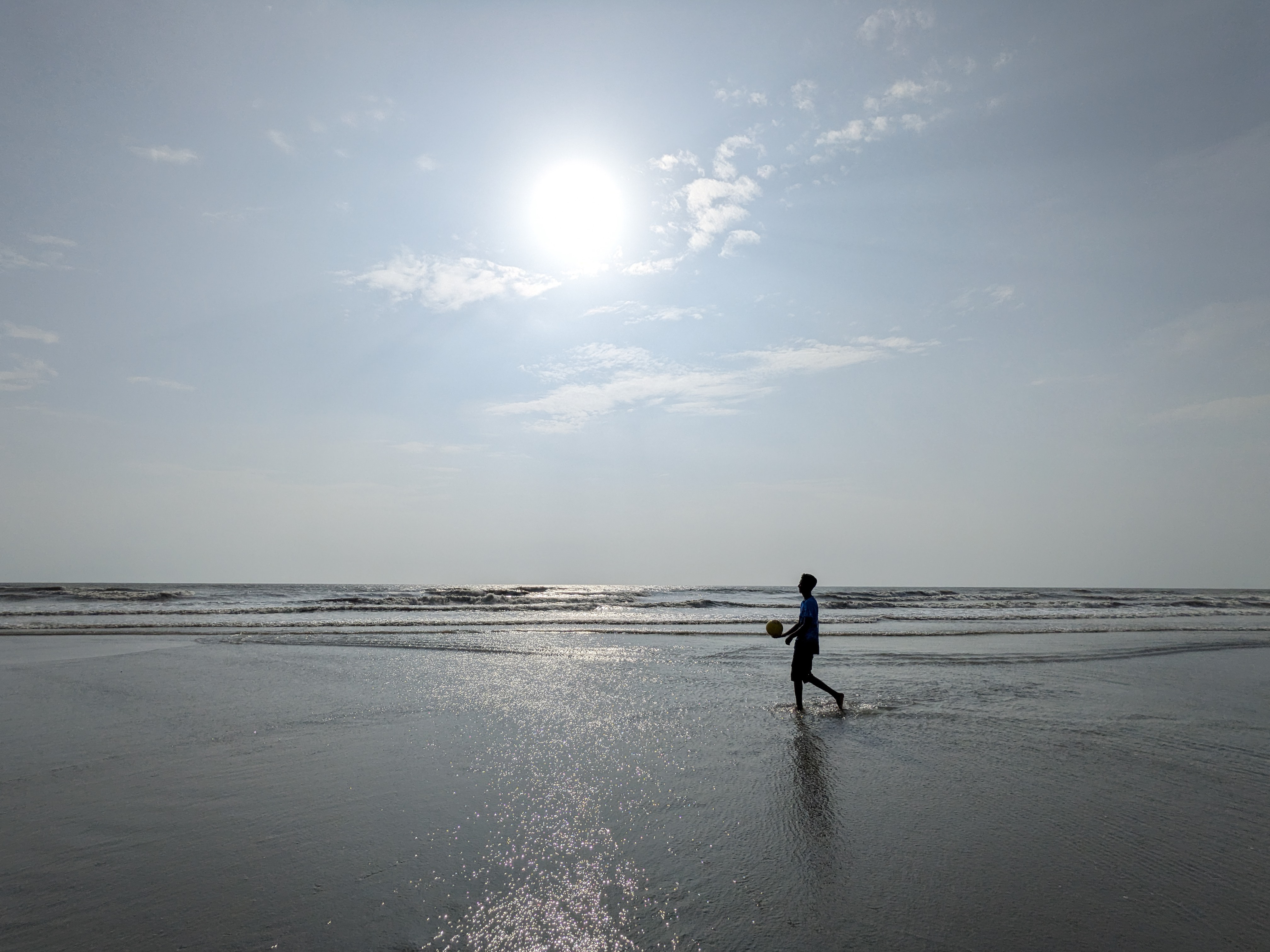 A bright sunny moment at Lal Kakra Beach, Cox’s Bazar. The calm sea glistens under the golden sunlight as a boy walks along the shore with a ball, capturing the simple joy and peaceful rhythm of coastal life.