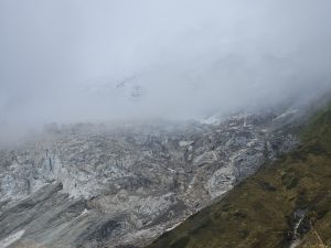 A mountainous landscape shrouded in clouds and mist. The rocky terrain features jagged, gray cliffs with patches of ice or snow visible, while the lower region shows grass and earthy tones. The overall atmosphere is moody and subdued, with the clouds creating a sense of mystery.
