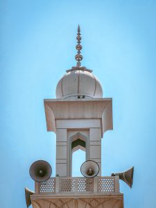 Close-up view of a mosque minaret with loudspeakers and a decorative dome against a clear blue sky