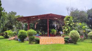 A large open-air pavilion with a red metal frame and sloped roof stands in a well-kept garden surrounded by trimmed grass, ornamental shrubs, and small palm trees