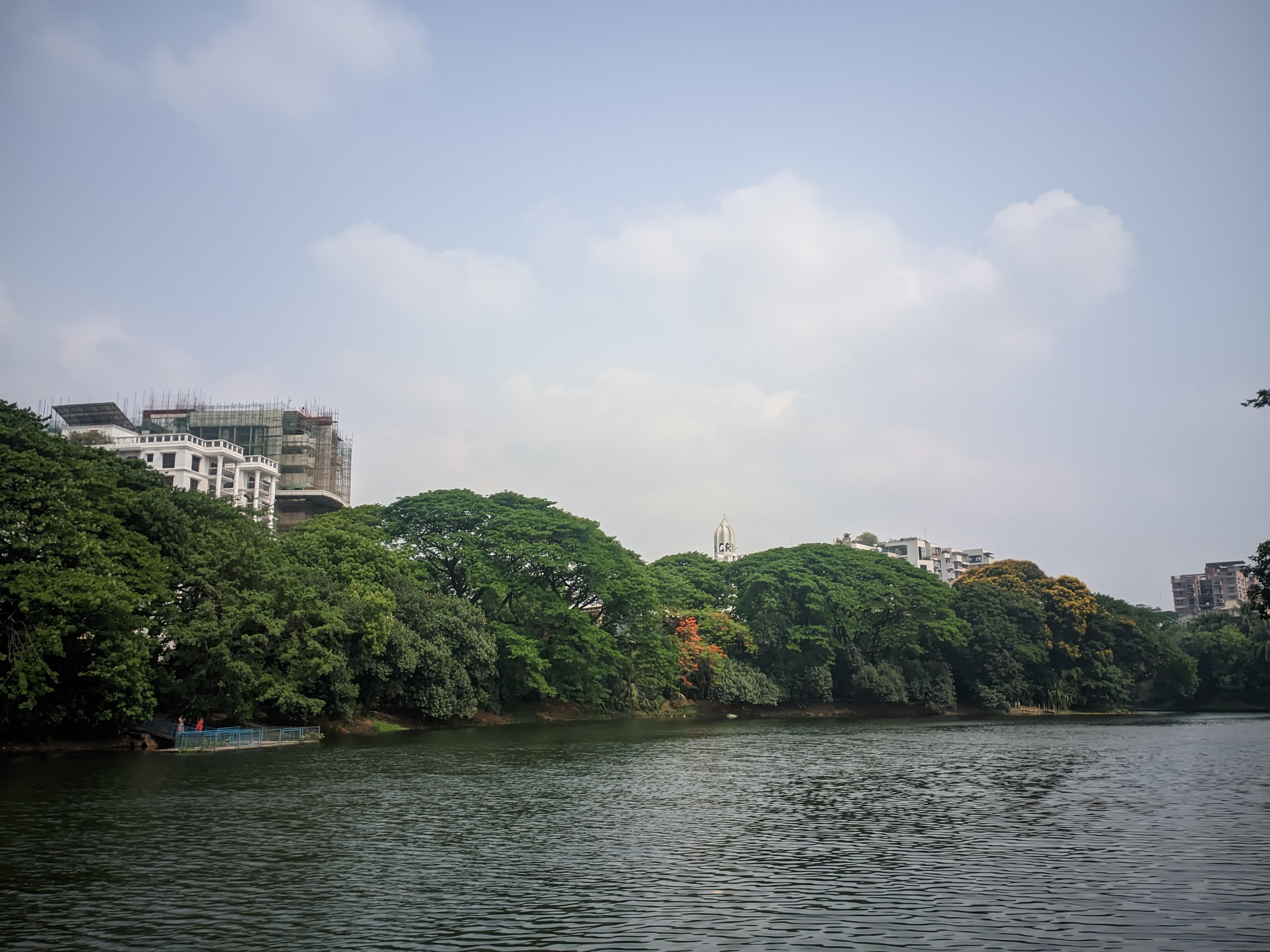 A calm river bordered by lush green trees, with buildings and a partly cloudy sky in the background. A construction site on the left hints at ongoing urban development.