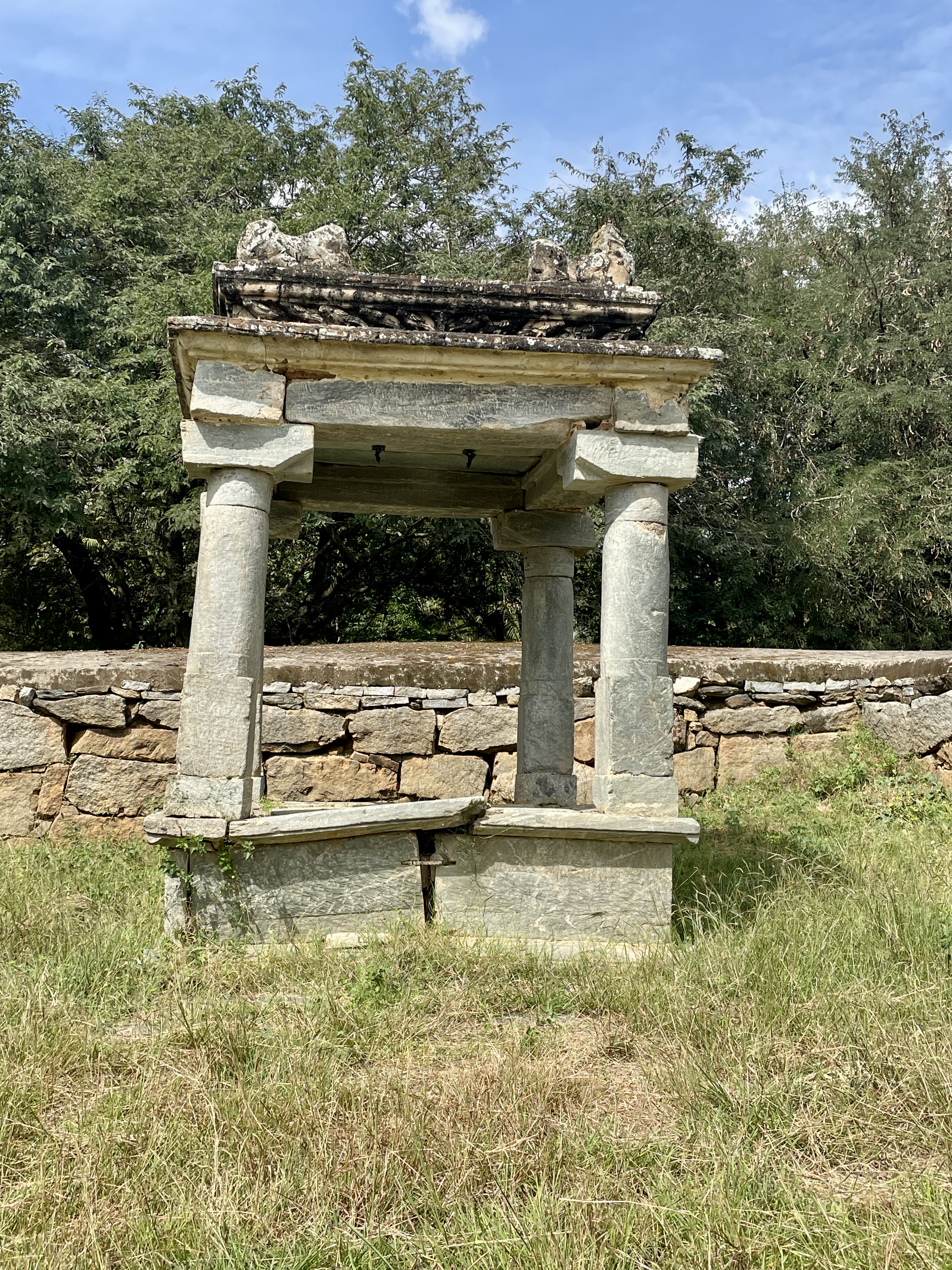 A small ruined stone mandapa (pavilion) with four pillars stands in a grassy patch beside the Lakshmi Narasimha Temple, Nuggehalli, Hassan. The structure shows signs of age but remains visually striking.