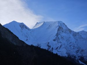 A dramatic mountain landscape featuring snow-covered peaks and rugged terrain, set against a clear blue sky with wispy clouds.