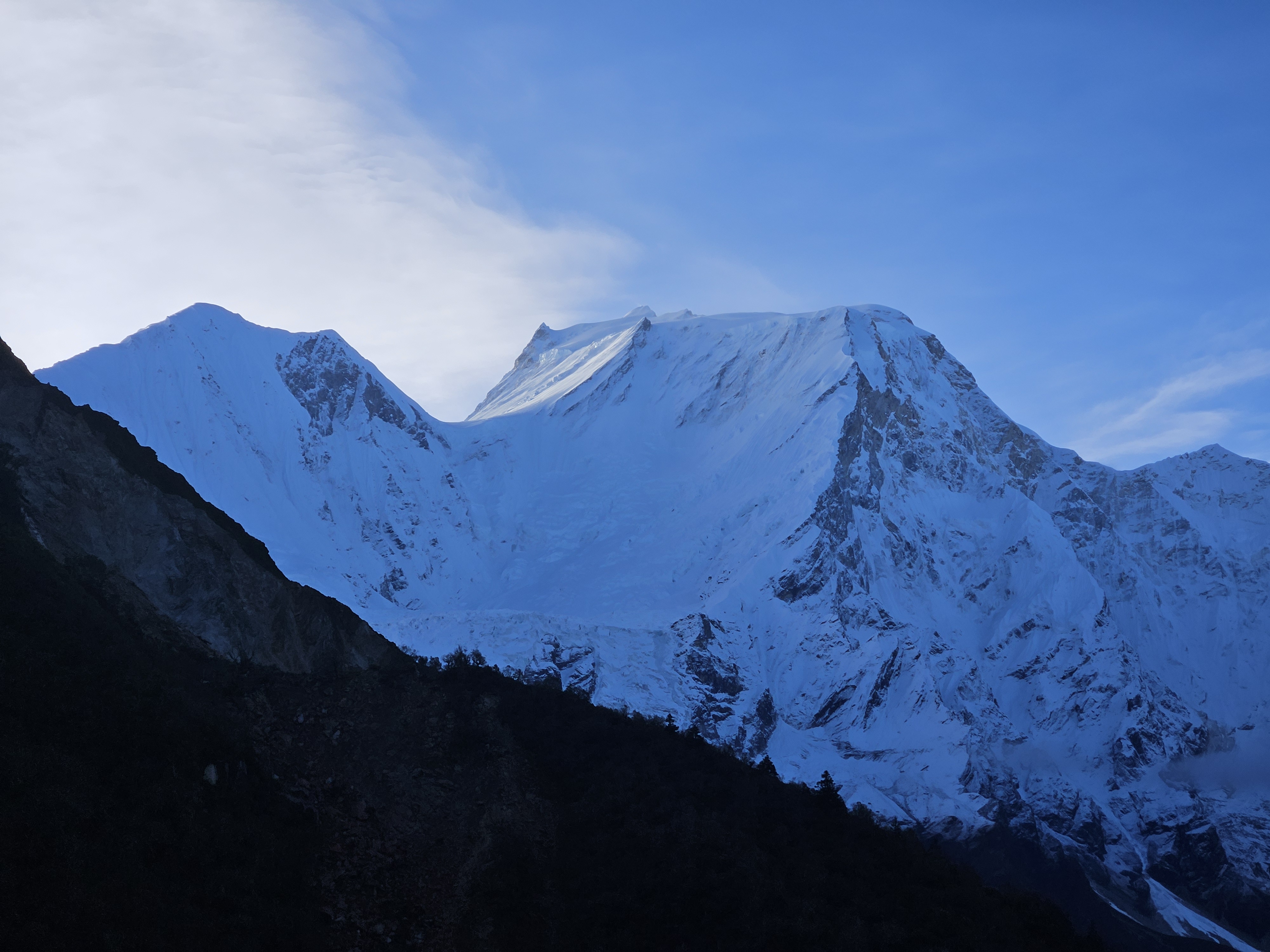A dramatic mountain landscape featuring snow-covered peaks and rugged terrain, set against a clear blue sky with wispy clouds.