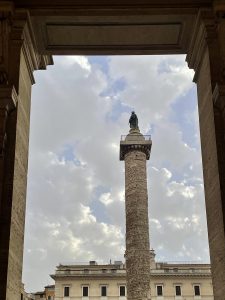 A view of the Column of Marcus Aurelius in Piazza Colonna, Rome, Italy, framed symmetrically between two stone pillars. The tall, intricately carved column stands against a soft, cloud-filled sky, with a statue of Marcus Aurelius on top and classic Roman architecture in the background. 