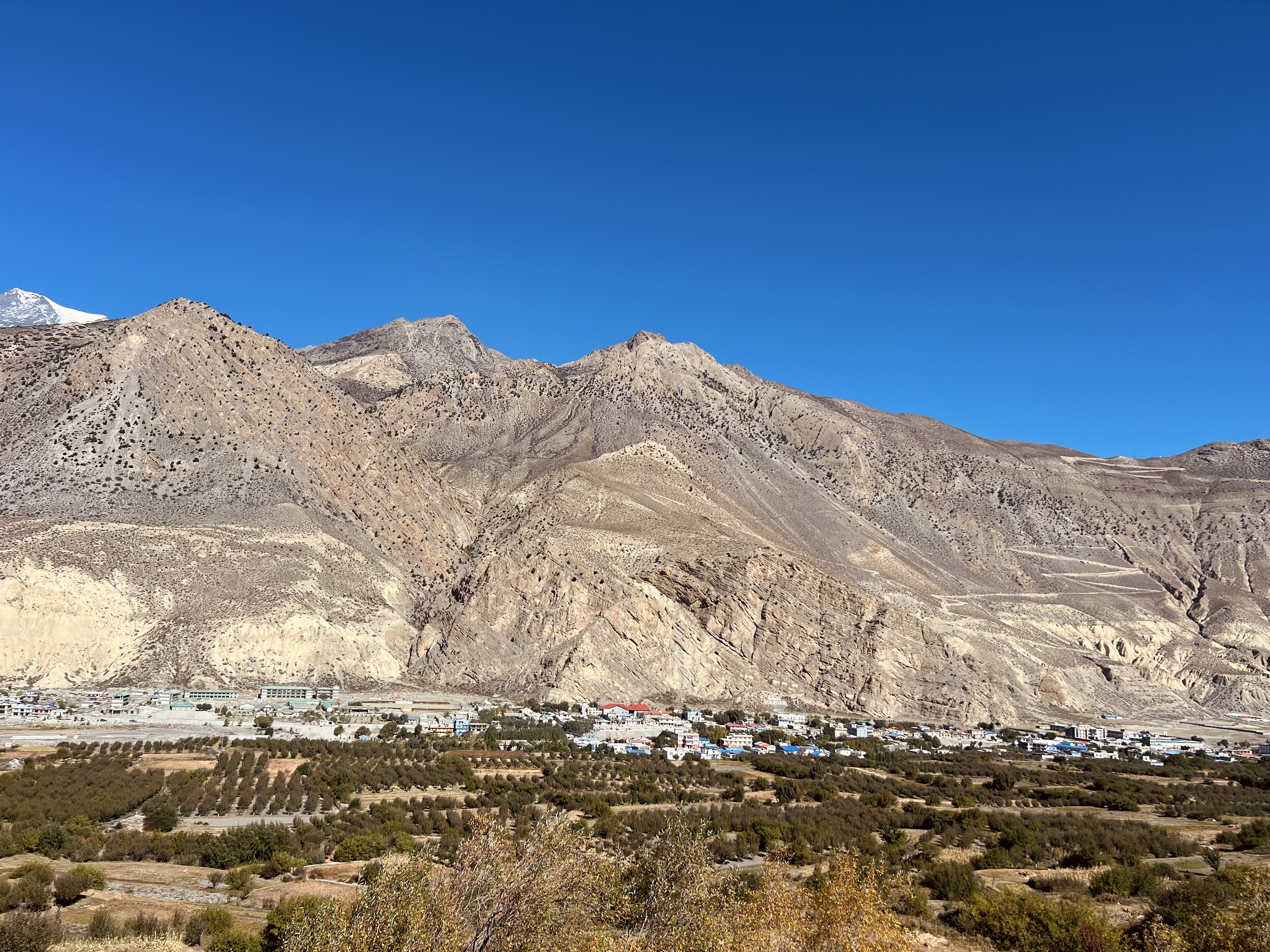Jomsom village features a scenic view of rugged, dry hills stretching across the landscape, set beneath a vast, clear blue sky.