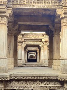 A view into the Adalaj Stepwell in Gandhinagar, Gujarat, showcasing its intricate Solanki-style architecture with multiple levels of carved stone pillars and arches. The symmetrical pillars create a vanishing corridor effect, highlighting the ornate carvings and perfect geometric balance.