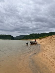 Jadukata River, Sunamganj. Raw, cloudy scene of a wooden boat on a golden-sandy bank. Two men are near the murky water. Dark green, forested hills rise in the distance under a heavy, grey sky