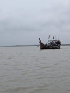 A wooden fishing boat with red-and-black flags floats on calm, grayish water under an overcast sky. Several people are visible on the boat, which sits slightly to the right of the frame, with distant land faintly visible along the horizon.