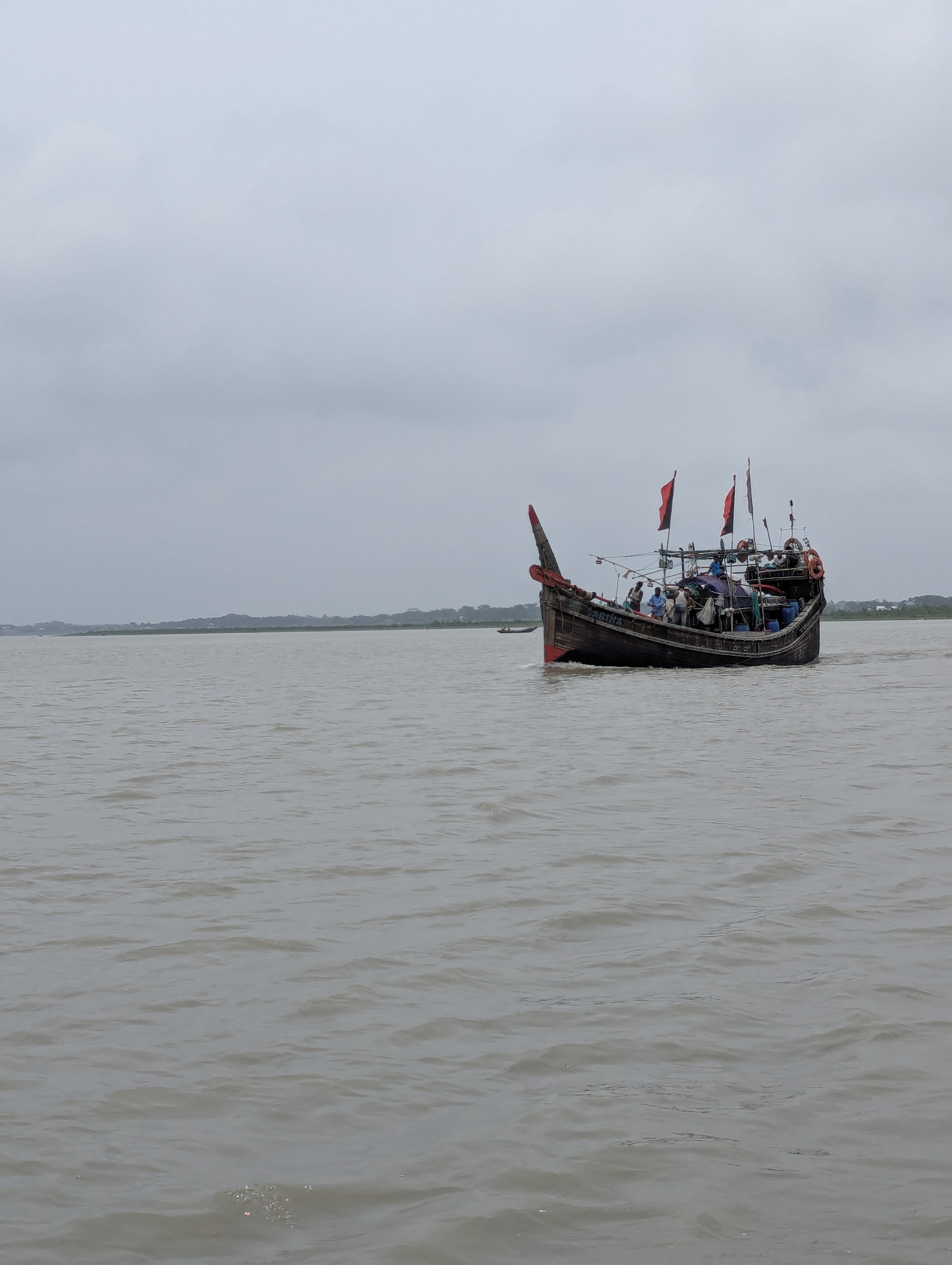 A wooden fishing boat with red-and-black flags floats on calm, grayish water under an overcast sky. Several people are visible on the boat, which sits slightly to the right of the frame, with distant land faintly visible along the horizon.