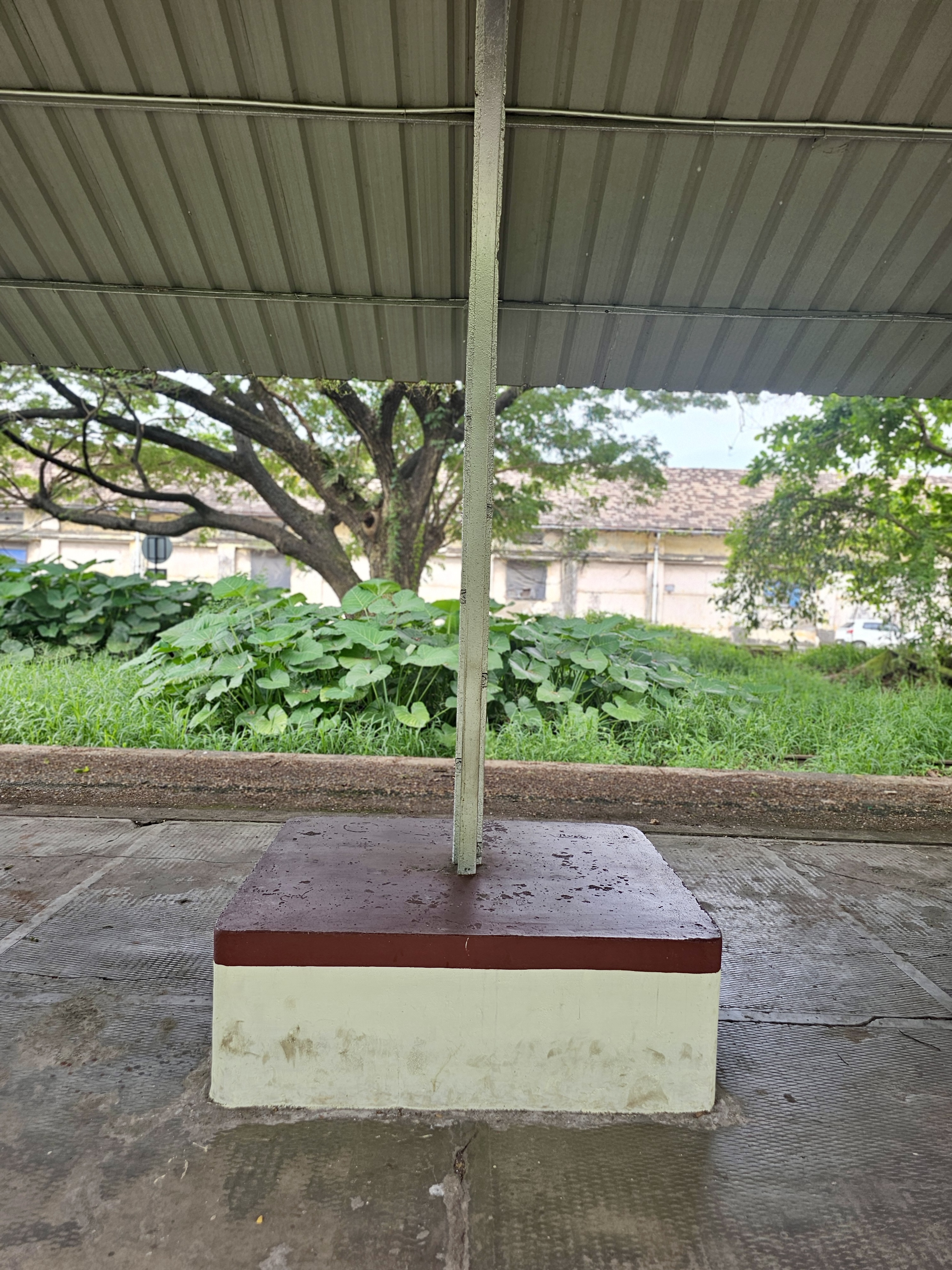 A close-up view of the metal pillar base supporting the shelter at the old Cochin Harbour Terminus railway station in Kochi. The platform is surrounded by greenery and unused tracks.