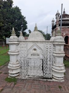 An ornate white tomb with domed spires stands amid greenery, with an ancient brick temple in the background.