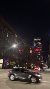 A night scene of an intersection in Chicago.  In the foreground is a car, slightly blurred by motion as it goes through the intersection. 