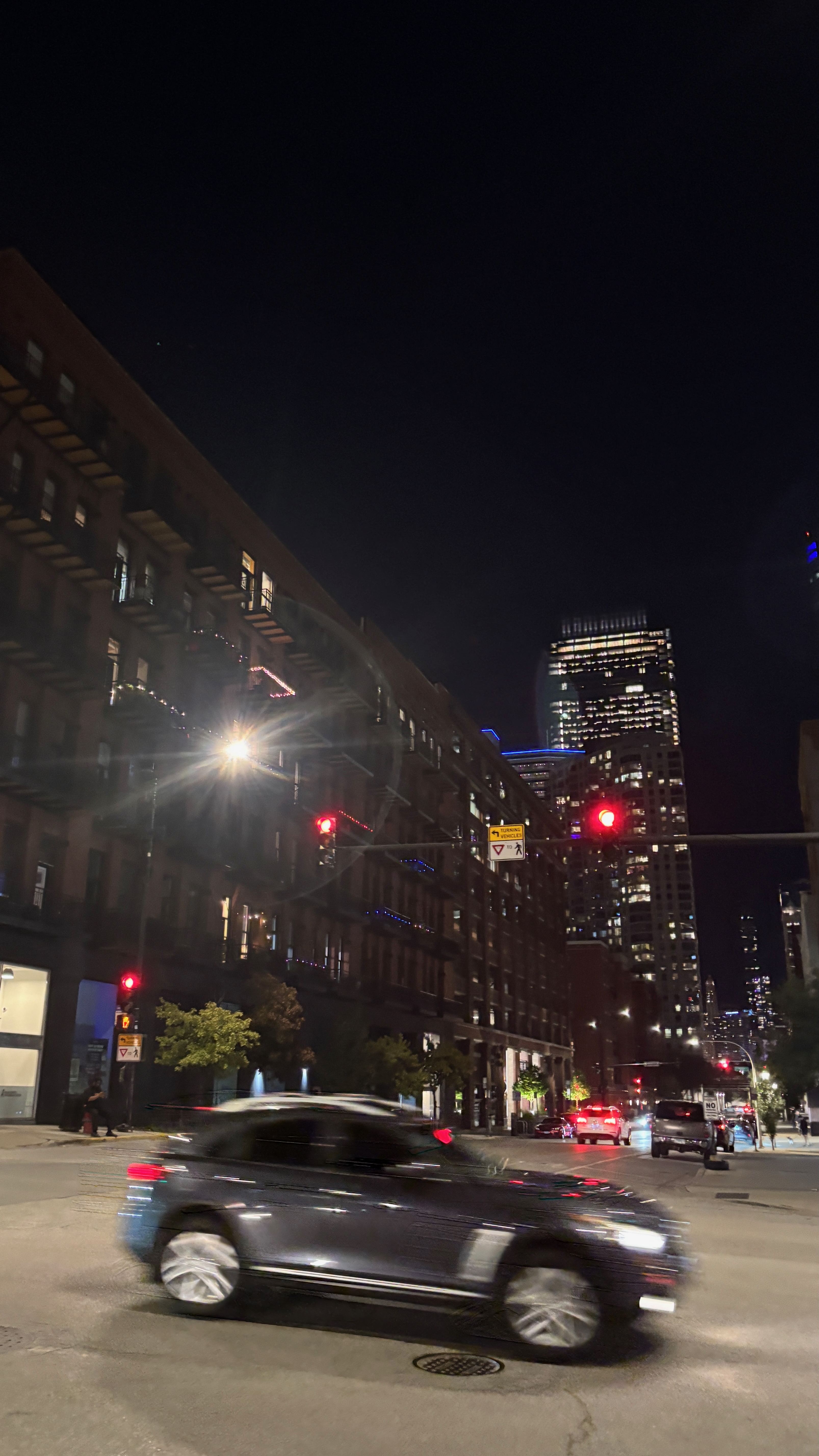 A night scene of an intersection in Chicago. In the foreground is a car, slightly blurred by motion as it goes through the intersection.