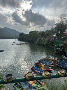 A peaceful lakeside landscape showing colorful wooden boats along the shore and a few floating on calm water surrounded by green hills and mountains.