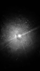 An image of a candle light taken from above illuminating a central circular spot surrounded by flared light showing a textured floor in a dark room.