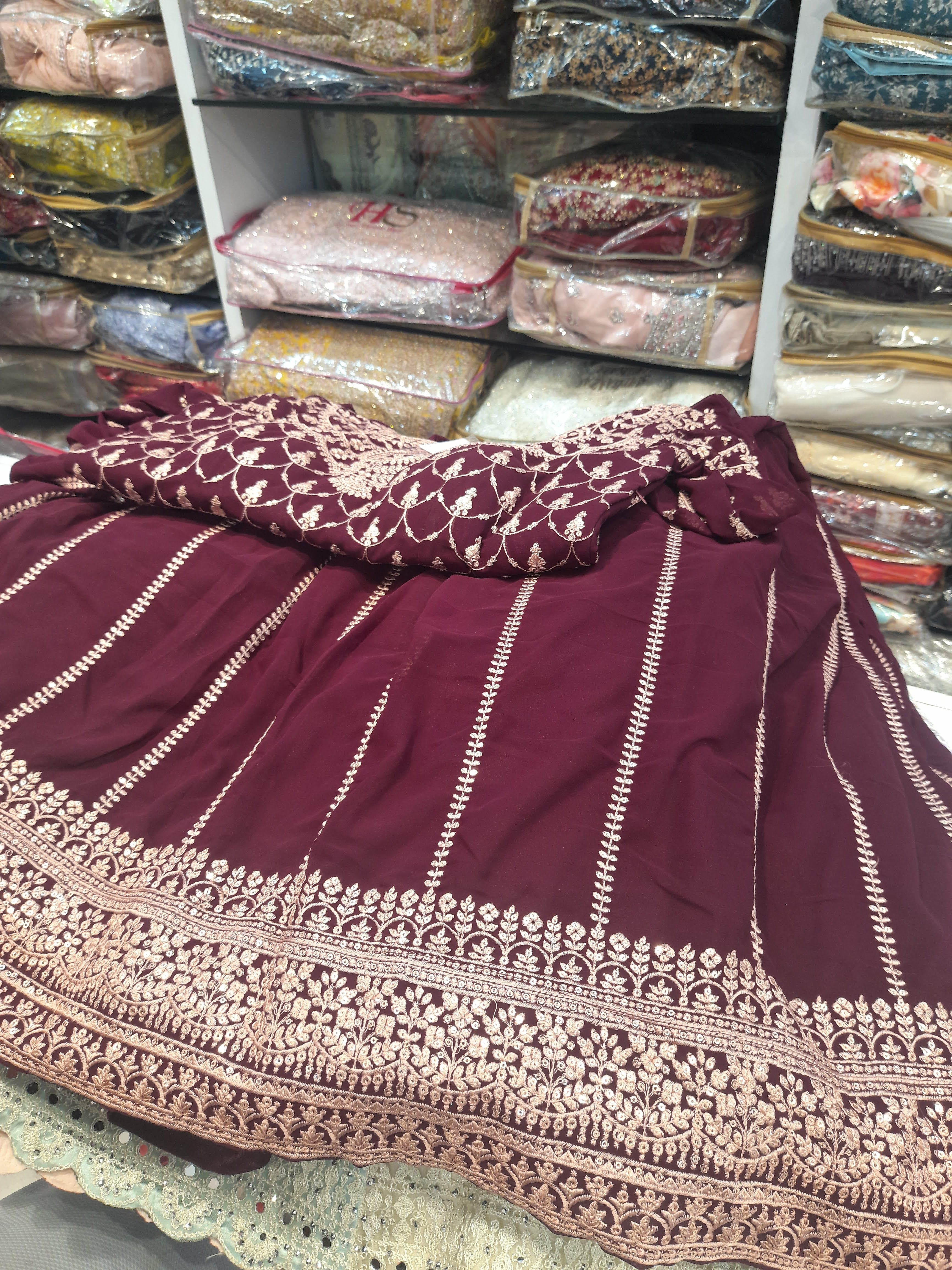 Rich maroon fabric with intricate white embroidery, displayed in a store. Shelves in the background hold folded, patterned textiles in clear bags, from kolkata, india.