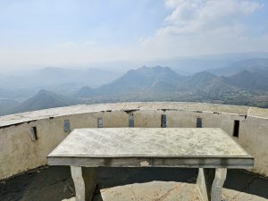 A panoramic view from Sajjangarh Palace in Udaipur, showing a stone table and wall in the foreground, with a stunning landscape of rolling hills and a valley under a bright, clear sky in the background. 