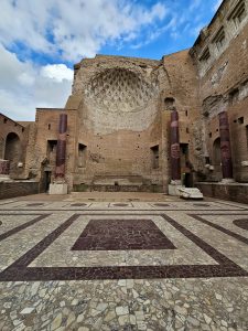 An ancient marble floor featuring red and white geometric patterns leads to a giant, curved wall with deep textures and arches in the Roman Forum of Rome, Italy.